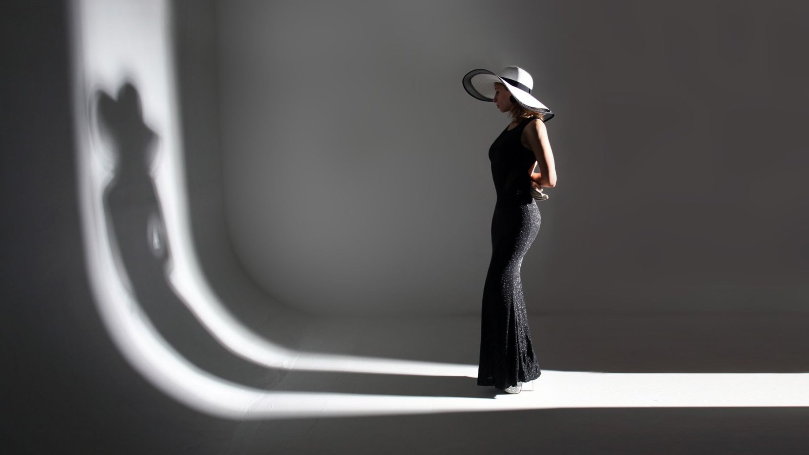 Full body of unrecognizable slim woman model in stylish long black dress and hat standing in dark studio and illuminated by light