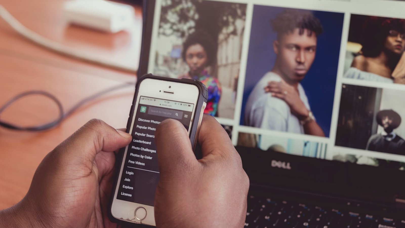 Close-up of hands holding smartphone in front of a laptop displaying images, indoors.