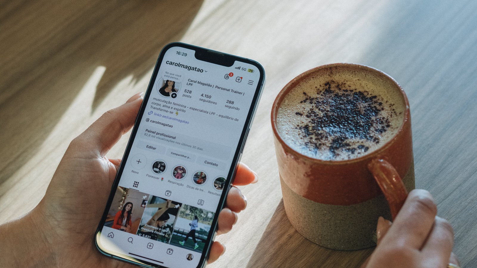 A hand holding a phone showing an Instagram profile grid of square thumbnails beside a coffee cup, the feed layout every platform size has to survive