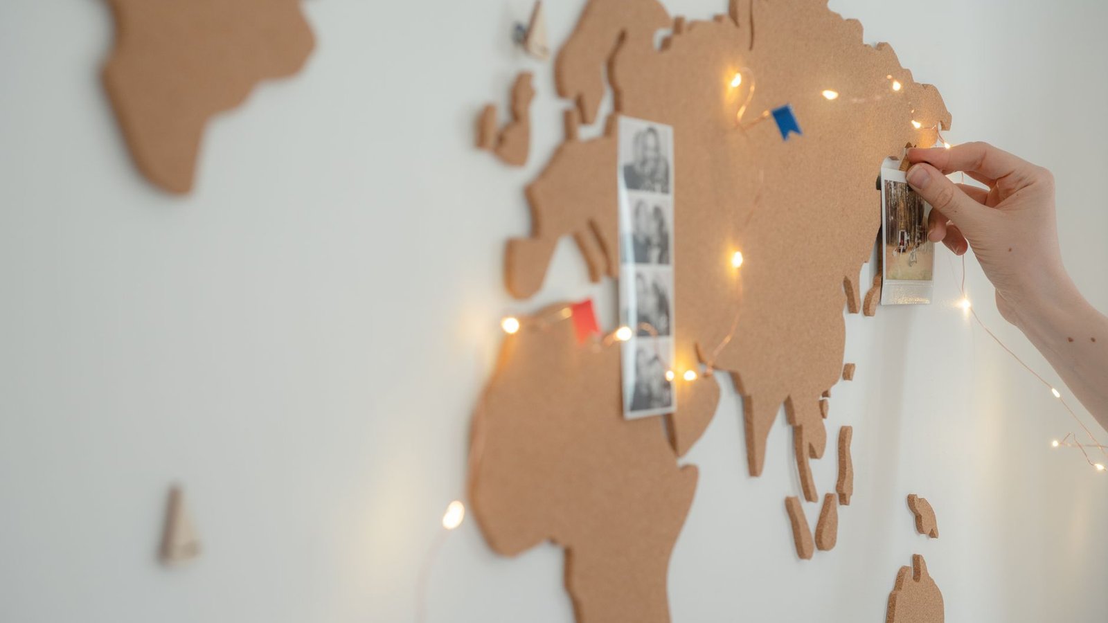 A hand pinning a small photo onto a cork world map strung with fairy lights, images collected onto a board the way a Pinterest board grows